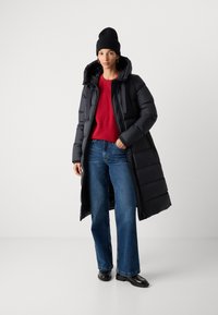 Vrouw in een lange zwarte pufferjack, zwarte beanie, rode trui, blauwe spijkerbroek en zwarte schoenen, staand met één hand in de zak.