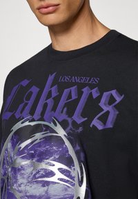 Sort T-shirt med lilla tekst "Los Angeles Lakers" og et sølv grafisk design med et svungen mønster på forsiden.