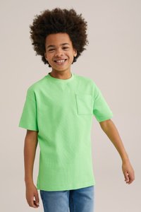 Gaiši zaļš īsām piedurknēm t-krekls ar apaļu kakla izgriezumu un krūšu kabatu, izgatavots no strukturētas auduma, valkāts ar zilām džinsēm.