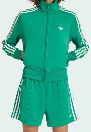 Persoon draagt een groene Adidas trainingsjas en bijpassende korte broek met witte strepen en logo, handen in de jaszakken.