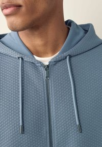Chaqueta con cremallera azul claro, con un patrón de diamante texturizado, cordones y una capucha suave. Detalle de cremallera metálica visible en el frente.