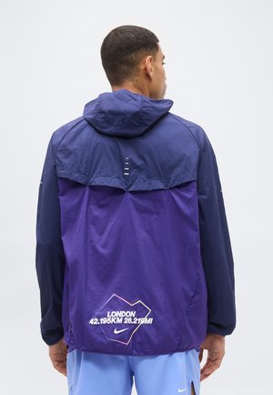 Άνδρας που φοράει μωβ και ναυτικό μπλε μπουφάν με κουκούλα με την επιγραφή "London 42.195KM 26.219MI" και το λογότυπο της Nike στην πλάτη, με την πλάτη γυρισμένη.