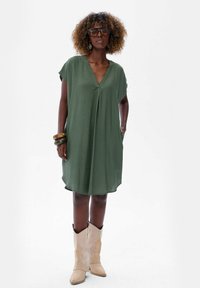 Zielona, sięgająca do kolan sukienka z lekkiego materiału, z dekoltem w kształcie litery V i krótkimi rękawami. Połączona z beżowymi kowbojkami i warstwami bransoletek.