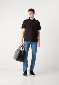 Svart polo skjorte, blå jeans, svarte joggesko med beige såler, holder en svart lærveske med doble håndtak og minimal maskinvare.