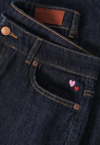 Temno modre kavbojke z oranžnim všitkom. Žep krasijo majhna rožnata in rdeča izvezena srca. Rjavo usnjeno etiketo krasi napis "TNTUUM."