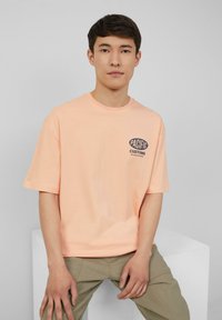 Licht perzikkleurig t-shirt met korte mouwen en een ronde hals. Voorzien van een marineblauw grafisch logo "PACIFIC CUSTOMS" op de linkerkant van de borst.