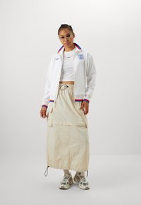 Hvit bomberjakke med rød og blå krage, logo på brystet. Hvit crop-top, beige midiskjørt med lommer, elastisk kant, parret med mønstrede sneakers.
