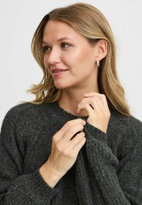 Dunkelgrauer, gestrickter Pullover mit gerippter Textur, runder Halslinie und etwas lockerer Passform. Das Model hält sanft den Ärmel.