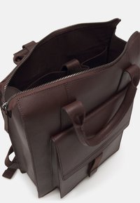 Sac à dos en cuir marron foncé avec fermeture éclair supérieure ouverte, poignées rembourrées, poche avant avec rabat et bretelles ajustables visibles.