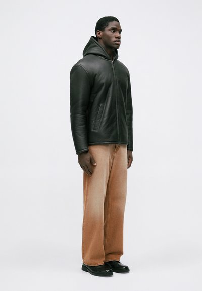 Schwarze Lederjacke mit Kapuze und glatter Textur, kombiniert mit hellbraunen weit geschnittenen Jeans und schwarzen Schuhen. Der Stil ist lässig und entspannt.