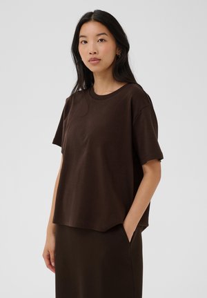 Jeune femme aux longs cheveux foncés portant un t-shirt ample marron foncé à manches courtes et une jupe assortie, debout avec une main dans la poche.
