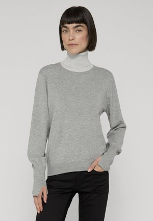 Vrouw met kort donker haar die een lichtgrijze coltrui onder een grijze trui met lange mouwen en zwarte broek draagt, staand met één hand in de zak.