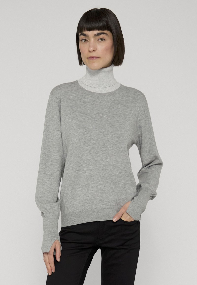 Femme aux cheveux courts et foncés portant un col roulé gris clair sous un pull gris à manches longues et un pantalon noir, debout avec une main dans la poche.
