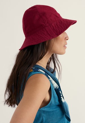 Mujer de cabello largo y oscuro, vistiendo un top azul sin mangas con flores y un sombrero rojo de cubo, mirando hacia la derecha contra un fondo liso.