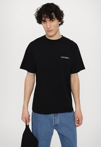 Svart bomull t-skjorte med korte ermer med teksten "No Problemo" i hvitt; kombinert med lyseblå denimjeans. Enkelt design og avslappet passform.