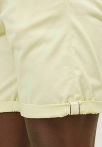 Světlé žluté šortky s měkkou texturou, s ohrnutým lemem a kontrastním červeným šitím. Detailní záběr na látku a design.