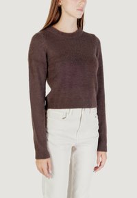 Donna che indossa un maglione aderente a maniche lunghe, peloso e marrone scuro, con pantaloni bianchi a vita alta, in piedi contro uno sfondo chiaro e semplice.