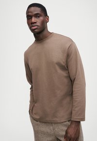 Smeđa košulja dugih rukava s okruglim izrezom, izrađena od mekog materijala. Ima opušteni kroj i minimalistički dizajn. Nosena je s teksturiranim hlačama.