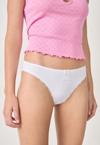 Mutande di cotone bianche con pizzo e un piccolo dettaglio a fiocco, con testo ricamato rosa "Divine". Indossate con una maglietta rosa con texture.