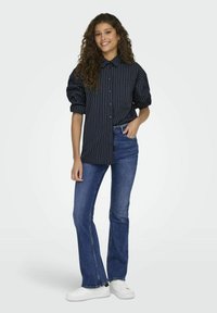 Marinrandig skjorta med knappar och puffärmar, matchad med utsvängda blå jeans och vita sneakers, på en ljus bakgrund.
