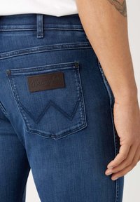 Jeans de mezclilla azul oscuro con corte recto, que presentan un parche de cuero marrón de Wrangler y un distintivo bordado de "W" en el bolsillo trasero.
