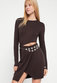 Hnedé dlhé šaty s prerezávaným detailom, majú zavinovací dizajn a strieborný opasok v tvare hviezdy. Hladká textúra látky.
