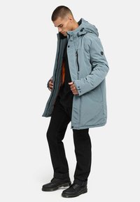 Ljusblå isolerad jacka med huva, dragkedja framtill och svart foder. Buren över en svart skjorta, tillsammans med mörka byxor och stövlar.