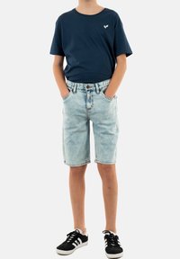 Dunkelblaues Baumwoll-T-Shirt mit einem weißen Logo, kombiniert mit hellblauen Jeansshorts und schwarzen Sneakers mit weißen Streifen. Sichtbare genähte Taschen.