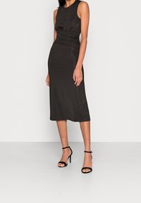 Schwarzes ärmelloses Midikleid mit gerafften Seitendetails und glatter Textur, kombiniert mit schwarzen hochhackigen Sandalen vor neutralem Hintergrund.