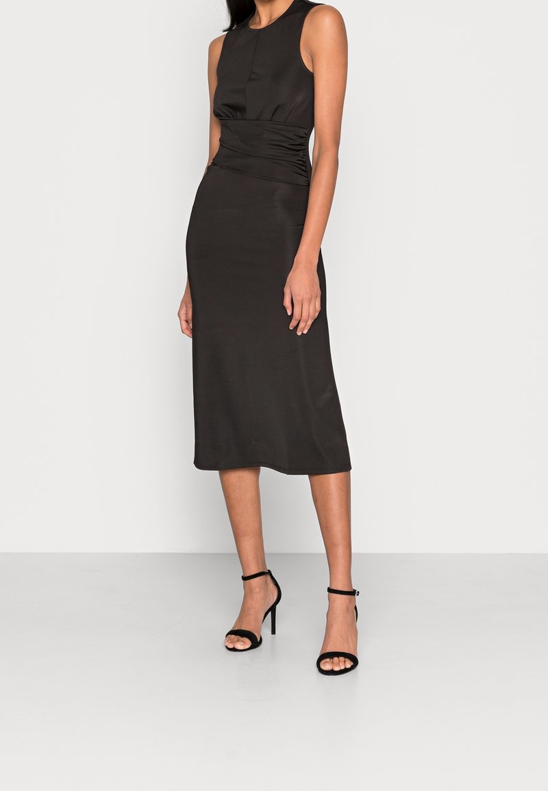 Schwarzes ärmelloses Midikleid mit gerafften Seitendetails und glatter Textur, kombiniert mit schwarzen hochhackigen Sandalen vor neutralem Hintergrund.