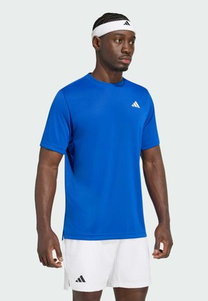 Atleet draagt een blauw Adidas T-shirt, witte korte broek en een witte Adidas hoofdband, staand tegen een effen achtergrond.