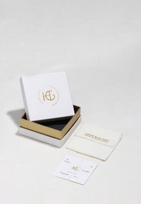 Bela škatla za nakit z zlatimi akcenti in logotipom, z črnim notranjim delom. Vključuje belo krpo za čiščenje in oznake, z zlatim besedilom.