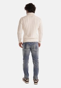 Maglione lavorato a maglia a treccia di colore crema con colletto alto, abbinato a pantaloni in denim azzurro chiaro strappati e scarpe da ginnastica marroni con dettagli bianchi.