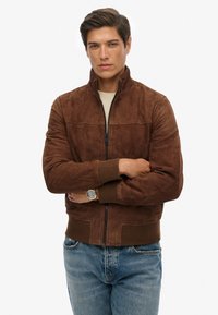 Giacca bomber in suede marrone con colletto alto e polsini a coste; presenta una zip frontale e cuciture tonali; abbinata a jeans blu.