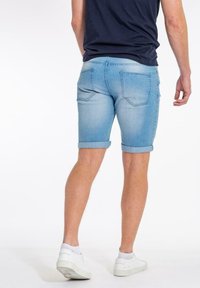 Henkilö, joka on pukeutunut käännettyihin vaaleansinisiin farkkushortseihin, tummansiniseen paitaan ja valkoisiin lenkkareihin, seisoo selkä kameraan päin valkoista taustaa vasten.