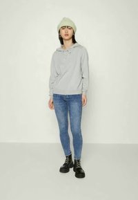 Uma hoodie cinza com cordão, jeans skinny azuis e botas pretas com atacadores. Um gorro verde claro completa o conjunto.