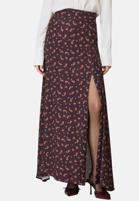 Gonna maxi in tessuto marrone scuro, decorata con piccoli motivi floreali rosa, con uno spacco laterale, abbinata a una blusa bianca e tacchi alti.