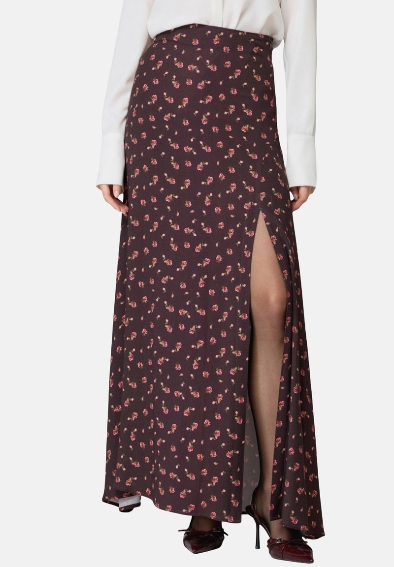 Gonna maxi in tessuto marrone scuro, decorata con piccoli motivi floreali rosa, con uno spacco laterale, abbinata a una blusa bianca e tacchi alti.