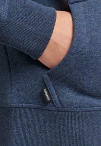 Sudadera de color azul marino con tela texturizada, puños acanalados, un bolsillo frontal y una pequeña etiqueta de marca negra.