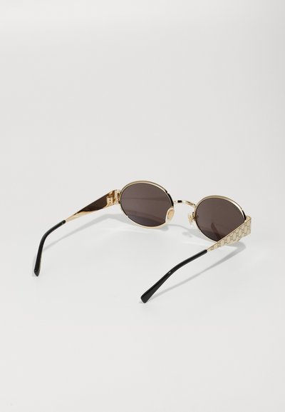 Gafas de sol ovaladas en tono dorado con lentes oscuros. Las patillas presentan un diseño texturizado, que se transforma a negro en las puntas.