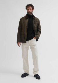 Brązowa kurtka z kordurowej tkaniny z brązowym kołnierzem, czarny golf, kremowe spodnie i czarne buty. Model stoi na jasnym tle.