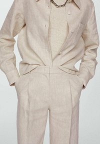 Chemise en lin beige clair avec une poche, assortie à un pantalon à coupe ajustée et pli visible. Accent de collier en chaîne argentée.