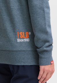 Grå sweatshirt med orange og hvid broderet tekst "!SLD Siden 1993" på venstre ærme og et rødt mærke ved manchetterne.