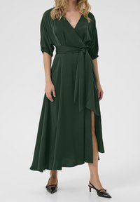 Lange, donkergroene wikkeljurk van lichte stof met een diepe V-hals, geplooide pofmouwen en een zijsplit. Gecombineerd met zwarte puntige schoenen.