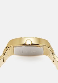 Montre-bracelet dorée avec un boîtier rectangulaire, un bracelet en métal et une couronne surélevée. L'arrière affiche des détails de marque et de modèle.