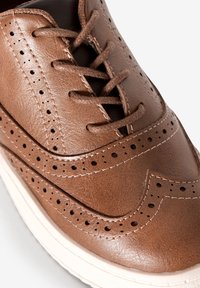 Chaussure en cuir marron avec des détails en brogue, des lacets et une finition texturée. La tige comporte des accents perforés et une semelle en caoutchouc blanc.