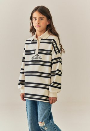 Pige med langt brunt hår iført en beige og sort stribet skjorte med krave og revne blå jeans, stående mod en ensfarvet lys baggrund.