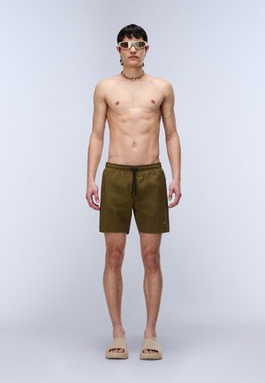 Mann, der olivgrüne Badeshorts, beige Sandalen, eine Perlenkette und weiße Sonnenbrille trägt, steht vor einem schlichten hellen Hintergrund.