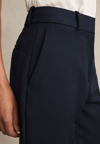 Prim-plan cu o persoană care poartă pantaloni bleumarin înalți pe talie, cu bucle pentru curea și buzunare laterale, asortați cu un top alb.