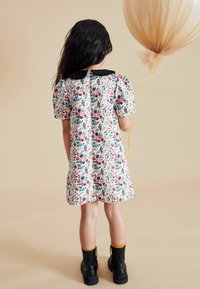 Robe florale avec des manches bouffantes courtes, col noir et fermeture à boutons. Associée à des bottes noires et un ballotin à pompons jaunes.